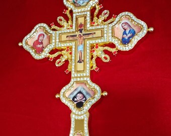Może przedstawiać: Ozdobny złoty krzyż z wbudowanymi wizerunkami religijnymi i błyszczącymi kryształowymi akcentami. Krzyż przedstawia centralne przedstawienie Jezusa na krzyżu, otoczone mniejszymi obrazami świętych i postaci religijnych. Na czerwonym tle.