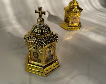 May include: Two gold-colored miniature church-shaped containers with ornate details and images of saints. The containers have a domed roof and a cross on top.