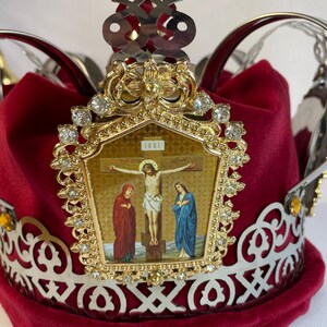 May include: A red velvet crown with a gold and silver filigree design. The crown features a gold emblem with a religious icon of Jesus on the cross, adorned with clear gemstones.