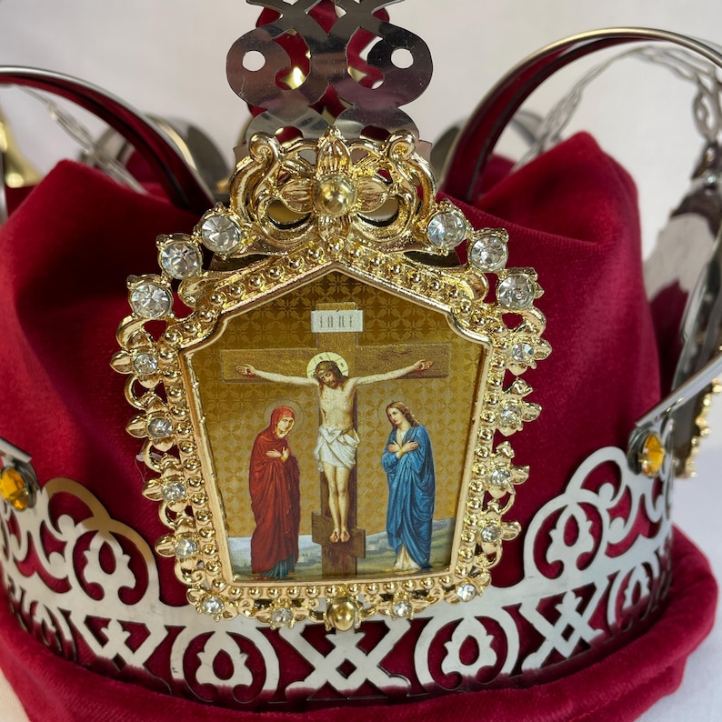 May include: A red velvet crown with a gold and silver filigree design. The crown features a gold emblem with a religious icon of Jesus on the cross, adorned with clear gemstones.