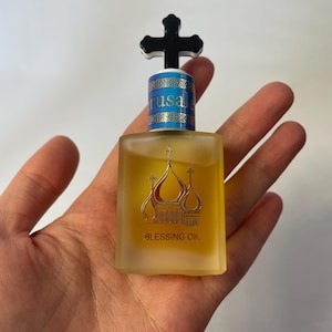 May include: A small, rectangular glass bottle of Blessing Oil, held in a hand. The bottle has a black cross-shaped stopper and a gold design of church domes. The label reads "Jerusalem" and "Blessing Oil".