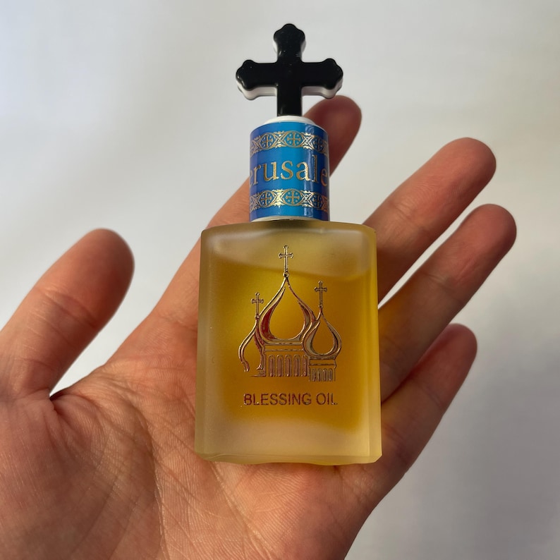 May include: A small, rectangular glass bottle of Blessing Oil, held in a hand. The bottle has a black cross-shaped stopper and a gold design of church domes. The label reads "Jerusalem" and "Blessing Oil".