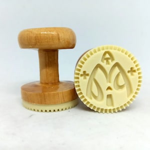 May include: Two wooden cookie stamps, one with a handle and one without. The stamp without a handle has a raised design of a crescent moon and stars.