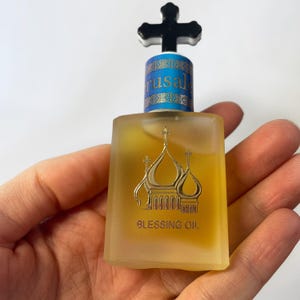 May include: A small, square glass bottle of Blessing Oil, held in a hand. The bottle has a gold label with a church design and the words "Blessing Oil". The cap is black with a cross on top, and the bottle has the word "Jerusalem" on it.