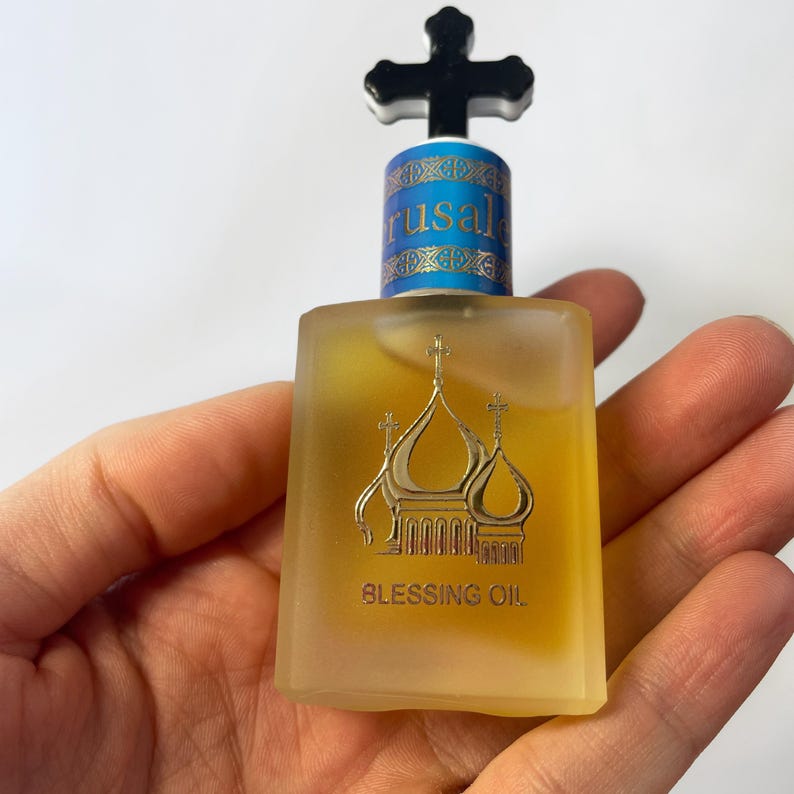 May include: A small, square glass bottle of Blessing Oil, held in a hand. The bottle has a gold label with a church design and the words "Blessing Oil". The cap is black with a cross on top, and the bottle has the word "Jerusalem" on it.
