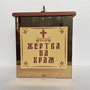Puede incluir: Una caja de donación rectangular de latón con tapa de madera. Una etiqueta crema en la parte delantera presenta una cruz y texto en color granate. La caja tiene un pequeño lazo de metal en la parte superior y un pequeño botón en la parte inferior.