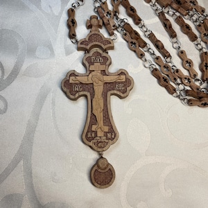 Puede incluir: Un colgante de cruz ortodoxa de madera con una imagen tallada de Jesucristo. La cruz es marrón con un borde marrón más oscuro y está unida a una cadena de cuentas de madera con eslabones de metal plateado. La cruz tiene texto religioso.