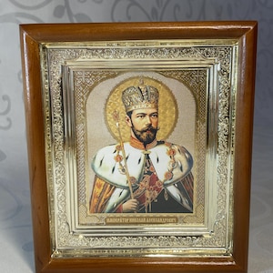 May include: A framed icon featuring a portrait of a man wearing a crown and holding a scepter. The portrait is surrounded by a gold-colored frame with ornate detailing. The frame is set within a wooden border.