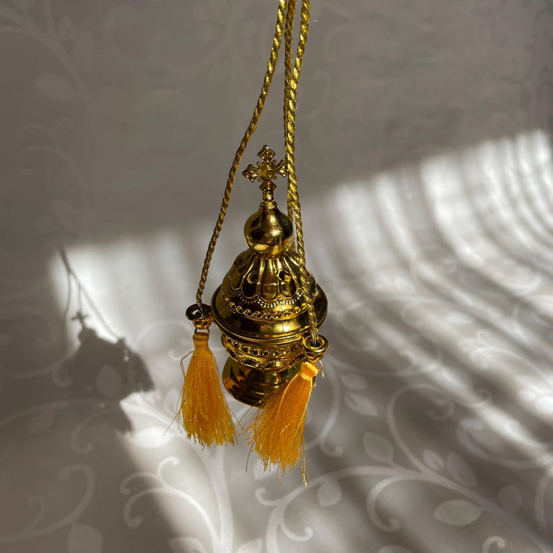 May include: A gold-colored censer with a cross on top and yellow tassels hanging from the bottom. The censer is suspended from a gold cord.