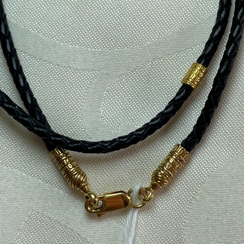 May include: A black leather cord necklace with gold accents. The necklace has a clasp closure and is braided.