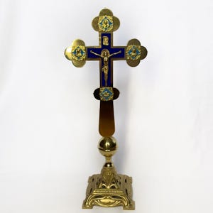 Op de afbeelding: Een goudkleurig kruis met blauwe emaille accenten en een figuur van Jezus Christus. Het kruis heeft sierlijke details en is gemonteerd op een decoratieve voet. Het kruis is ongeveer 30 cm hoog.