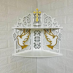 May include: White corner shelf with a decorative design featuring a gold cross and two doves with gold wings. The shelf has an ornate, cut-out design and is mounted on a white brick wall.