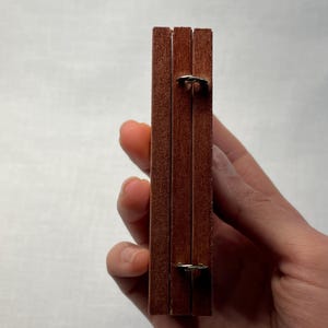 K&ouml;nnte beinhalten: Eine Holzkiste mit drei F&auml;chern, jedes mit einem Metallscharnier. Die Kiste ist geschlossen und wird in einer Hand gehalten.