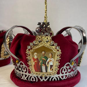 May include: A burgundy velvet crown with silver filigree and a gold emblem. The emblem features an icon of three figures and is adorned with clear crystals. A gold spire and a silver Om symbol are at the top.