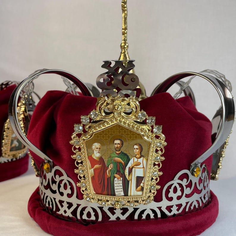 May include: A burgundy velvet crown with silver filigree and a gold emblem. The emblem features an icon of three figures and is adorned with clear crystals. A gold spire and a silver Om symbol are at the top.