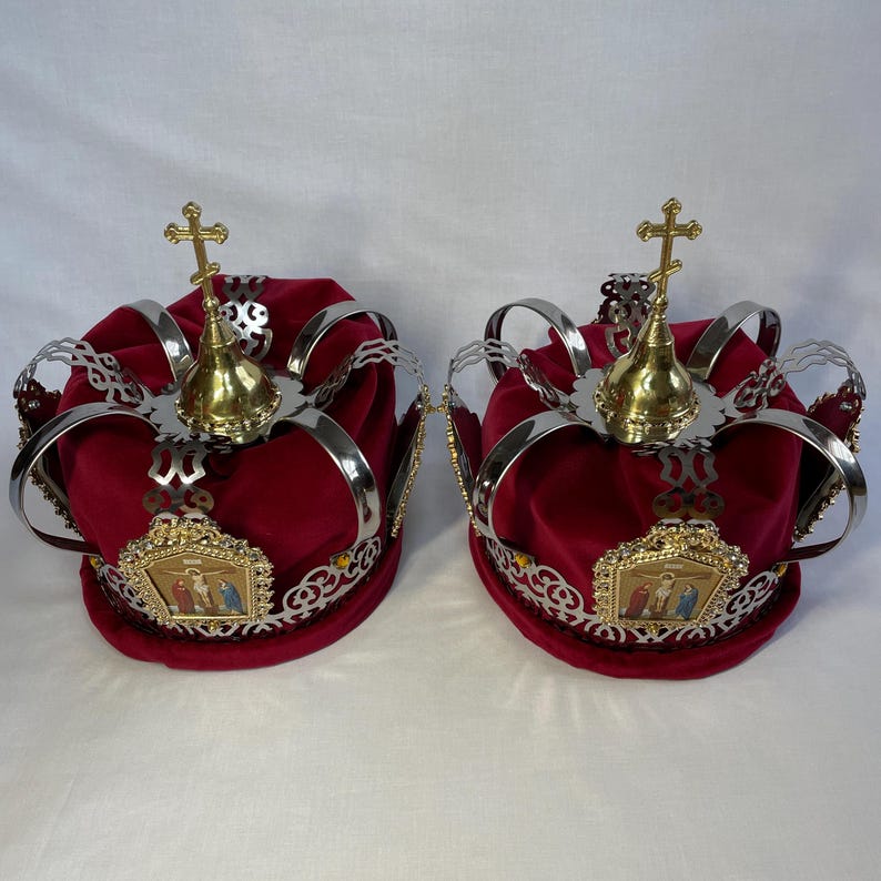 May include: Two ornate red velvet crowns with gold crosses and silver accents. Each crown features a gold emblem with a religious image and is trimmed with gold and silver filigree. The crowns are likely for a religious ceremony.