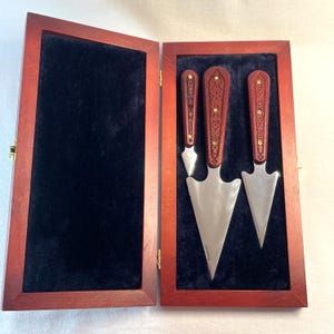 May include: A wooden box with a dark red finish, open to reveal three cheese knives. The knives have ornate, dark red handles with gold accents and stainless steel blades. The interior of the box is lined with black velvet.