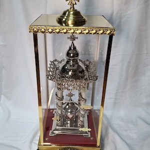 May include: A gold-toned metal stand with a glass case, featuring a silver-toned metal religious icon with intricate details. The stand has a red velvet base and ornate feet.