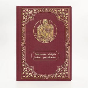 Pode incluir: Um livro bordô com detalhes decorativos dourados. A capa apresenta uma moldura dourada ornamentada com uma ilustração religiosa e texto. O título do livro está escrito em letras douradas. O livro está fechado e em pé.