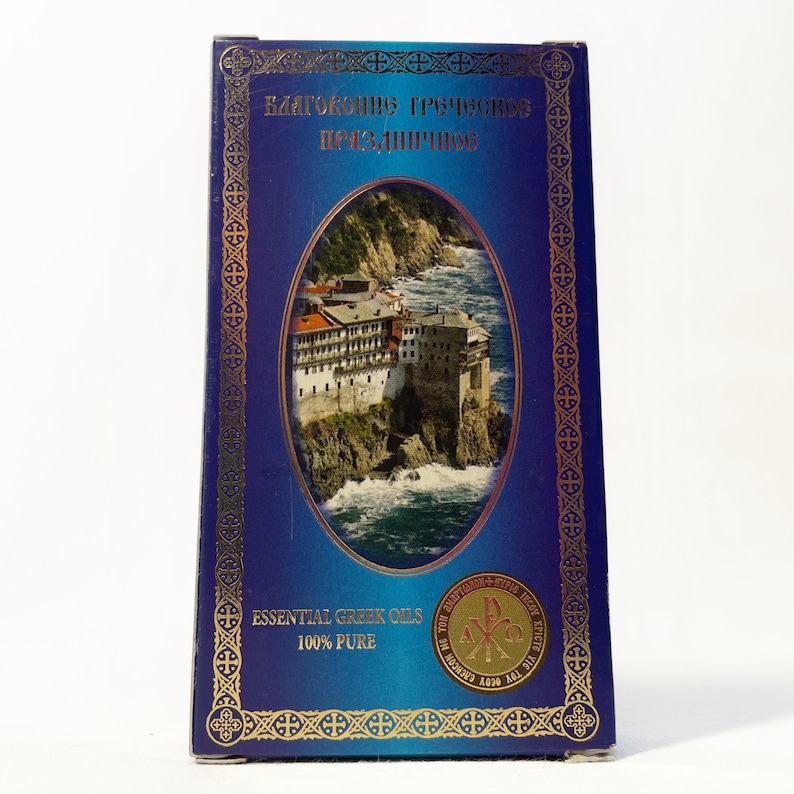 May include: A rectangular box with a dark blue background and gold trim. The front features an oval image of a building on a cliff overlooking the sea. Text reads "Essential Greek Oils 100% Pure".