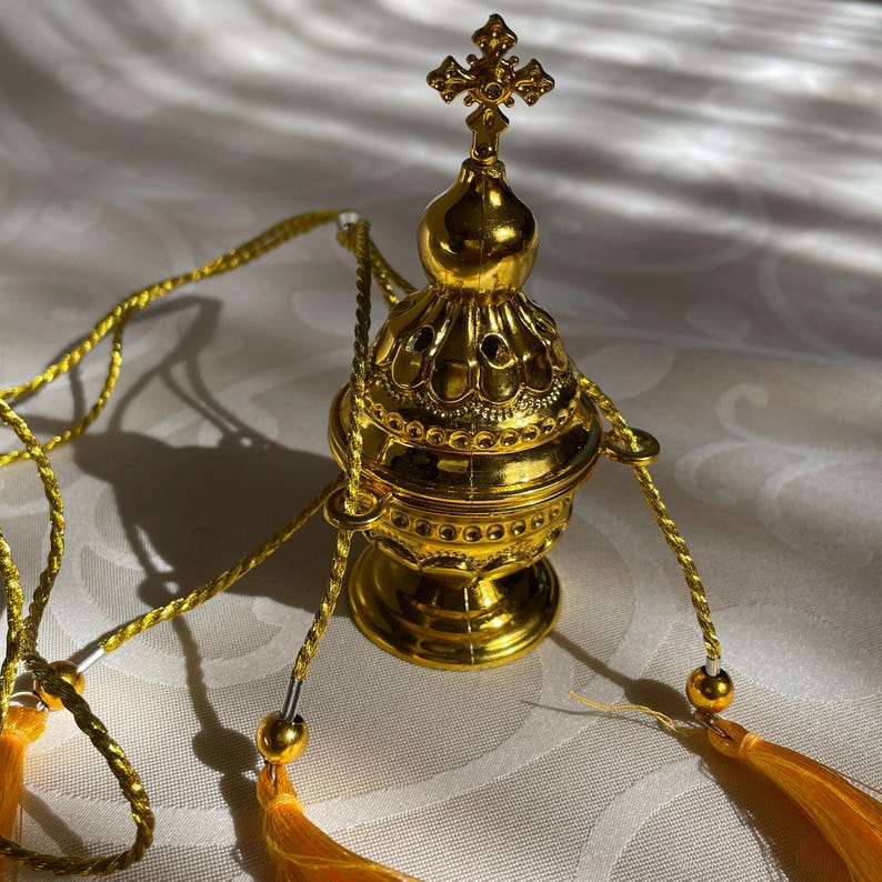 May include: A gold-colored metal censer with a cross on top and a chain with tassels. The censer is ornate and has a decorative design.