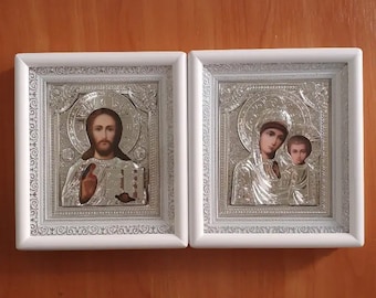 May include: Two white framed religious icons. The icon on the left depicts Jesus Christ with a golden halo. The icon on the right depicts the Virgin Mary holding baby Jesus.