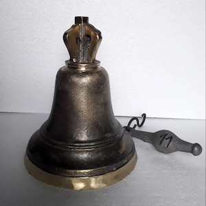 Puede incluir: Una campana de metal en tonos bronce oscuro con un borde dorado pulido y un asa decorativa en forma de corona en la parte superior. Un badajo de metal gris separado con el número 11 inscrito en el lateral está colocado junto a la campana.