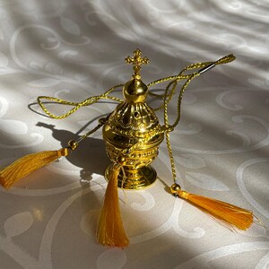 May include: A gold-colored metal censer with a cross on top and a chain with tassels. The censer is decorated with intricate details.