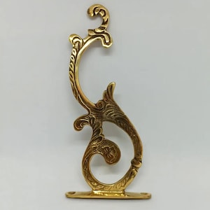 May include: A gold metal decorative bracket with a scrolled design. The bracket has a flat base with two holes and a decorative top with a scroll design.