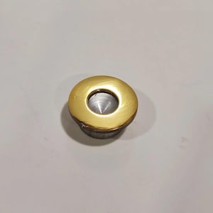 May include: A close-up of a brass-colored bottle stopper with a circular design. The stopper has a gold-colored top and a silver-colored base. The center of the stopper has a hole. The stopper is on a white surface.