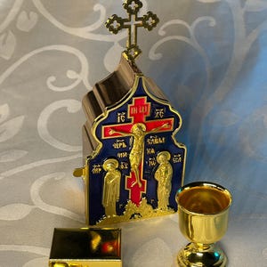Puede incluir: Un conjunto de iconos religiosos de color dorado y ornamentado con un diseño de esmalte azul y rojo. El icono representa una cruz con una figura de Jesucristo y dos figuras a cada lado. El conjunto también incluye un pequeño cáliz y una caja dorada.