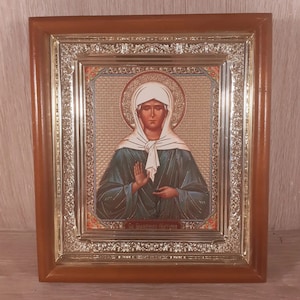 May include: A framed religious icon featuring a woman in a white head covering and blue robes. The icon has a gold halo and is set within an ornate gold and brown wooden frame. The image has text at the bottom.