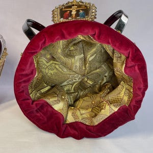 May include: A red velvet bag with a gold patterned interior. The bag has black handles and a decorative top with a religious scene. The bag's interior is a gold brocade fabric.