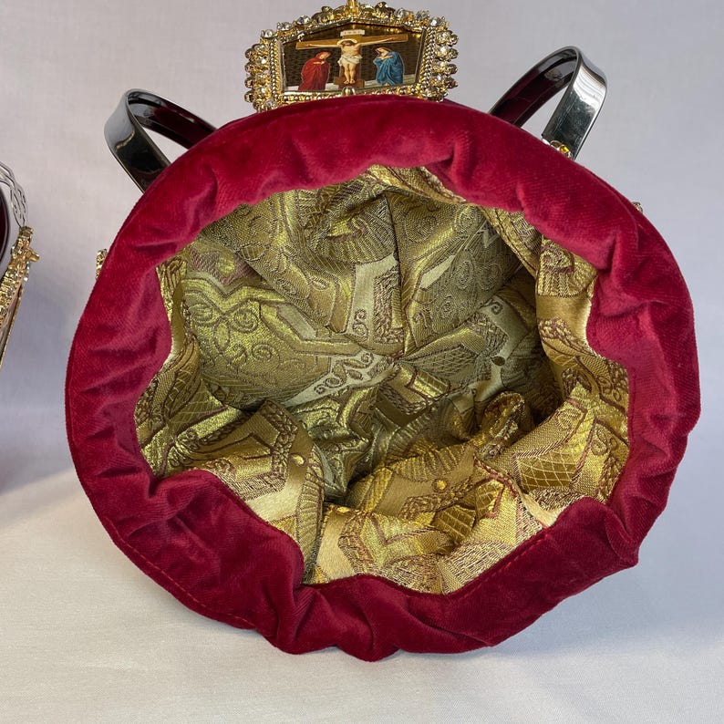 May include: A red velvet bag with a gold patterned interior. The bag has black handles and a decorative top with a religious scene. The bag's interior is a gold brocade fabric.