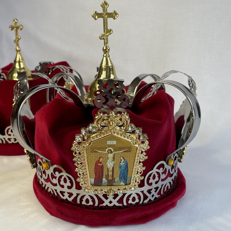 May include: Two ornate crowns with red velvet bases, silver filigree, and gold accents. Each crown features a gold cross and a religious icon depicting a crucifixion scene. The crowns are likely used for religious ceremonies.