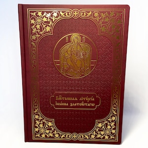 Könnte beinhalten: Ein weinrotes Buch mit goldenen, verzierten Designs und Text. Das Cover zeigt eine goldene Illustration einer religiösen Figur und Text in einer Fremdsprache. Der Einband des Buches hat ein strukturiertes Muster.