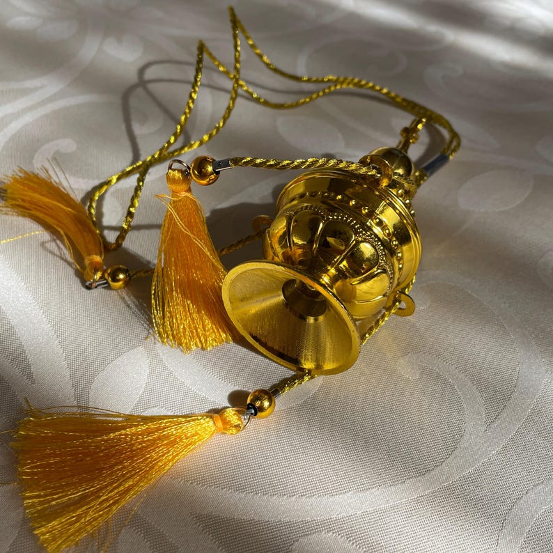 May include: A gold-colored metal incense burner with a decorative design and a tassel hanging from it. The burner is attached to a gold cord.