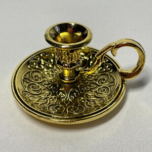 May include: A gold-colored candle holder with a round base and a handle. The base features an ornate, swirling design. The candle holder has a cup-shaped top to hold a candle.