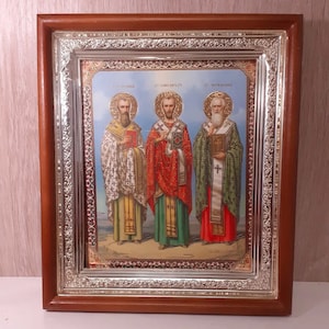 Op de afbeelding: Een religieus icoon in een lijst met drie figuren met halo's, elk met een boek of kruis. De figuren zijn gekleed in kleurrijke gewaden, tegen een blauwe luchtachtergrond. De lijst is bruin met een zilveren filigraanrand.