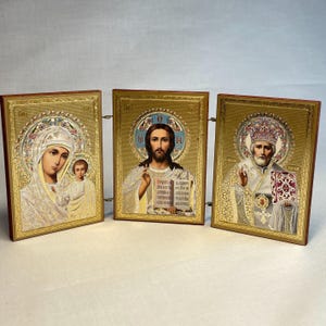 K&ouml;nnte beinhalten: Ein Satz von drei goldgerahmten religi&ouml;sen Ikonen. Die Ikonen zeigen die Jungfrau Maria und das Kind, Jesus Christus und einen Heiligen. Die Ikonen sind auf einem Holzst&auml;nder montiert.