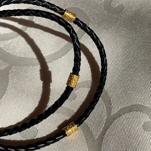 May include: Two black leather braided cords with gold cylindrical beads. The cords are intertwined and the beads are spaced evenly apart.