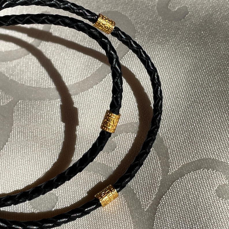 May include: Two black leather braided cords with gold cylindrical beads. The cords are intertwined and the beads are spaced evenly apart.