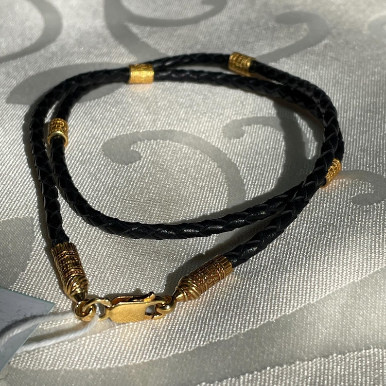 May include: A black leather cord necklace with gold accents. The cord is braided and has a gold clasp.