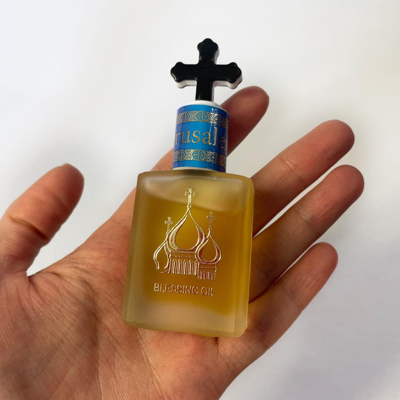 May include: A small, square bottle of Blessing Oil with a black cross-shaped stopper. The bottle has a gold-colored liquid inside and a gold illustration of a church. The word "Jerusalem" is printed on the blue and gold label.