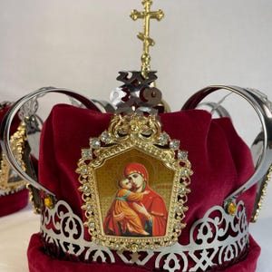 May include: A red velvet crown with silver accents and a gold cross. The crown features a gold-framed icon of Mary and Jesus, adorned with clear crystals. The crown is likely used for religious ceremonies.