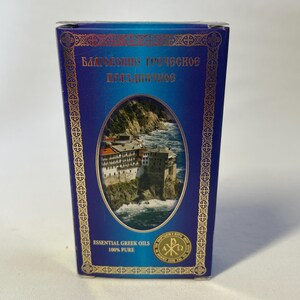 May include: A blue box with gold trim and text in a foreign language. The box features an oval image of a building on a rocky coast with waves. The text "ESSENTIAL GREEK OILS 100% PURE" is printed at the bottom.