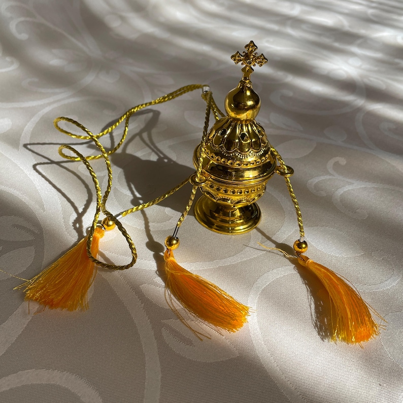 May include: A gold censer with a cross on top and three yellow tassels hanging from it. The censer is on a white surface.