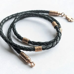 以下が含まれることがあります： 金色の金属製のアクセントが付いた黒い革ひもネックレス。ひもは編み込まれており、留め具が付いています。