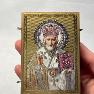 K&ouml;nnte beinhalten: Eine goldgerahmte Ikone, die eine religi&ouml;se Figur mit wei&szlig;em Bart und Krone darstellt. Die Figur h&auml;lt ein Buch und ein Kreuz. Die Ikone ist von einem dekorativen Rahmen umgeben.