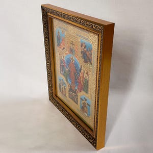 May include: A framed religious icon with a detailed depiction of biblical scenes. The artwork features a central figure surrounded by other figures, set against a gold background. The frame is a light brown color with a dark brown decorative border.